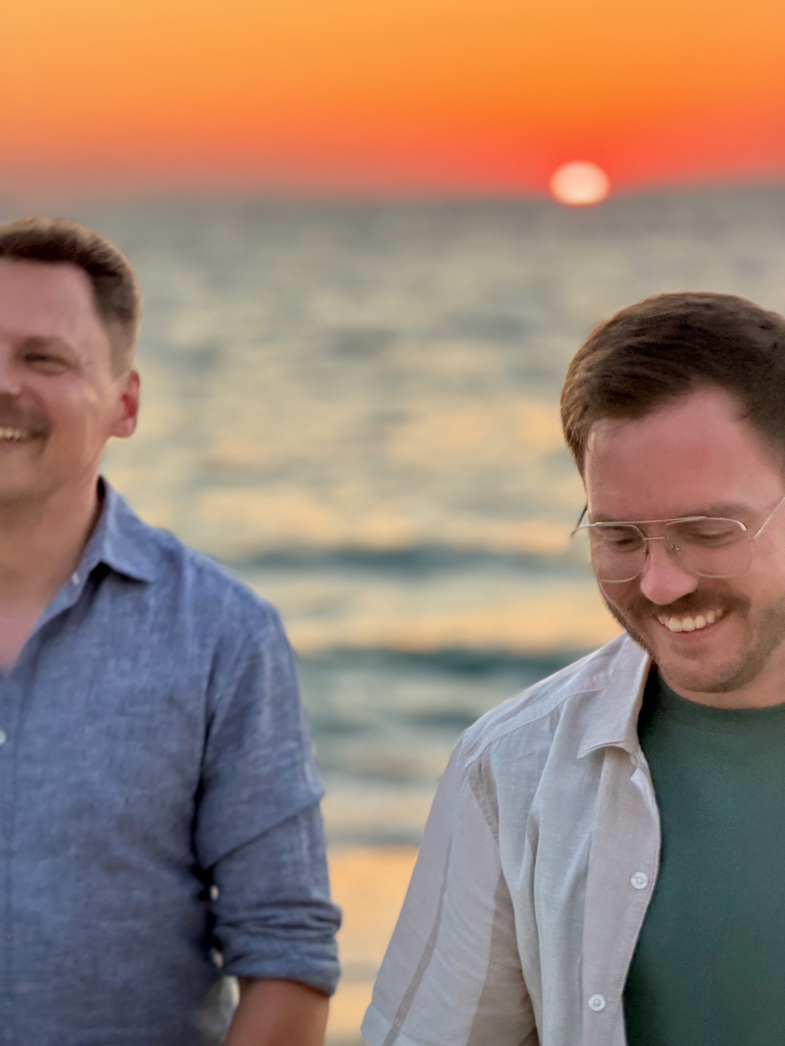 Stefan und Steffen lachen gemeinsam am Meer bei Sonnenuntergang.